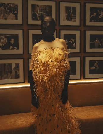 Adult female wearing a feather-covered gown and full face covering standing indoors with framed photos in the background.