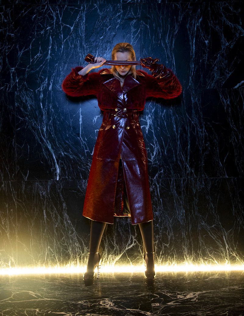 Stylish woman in a shiny red coat, knee-high boots, and sunglasses posing against a marble wall with neon lighting for a fashion shoot.