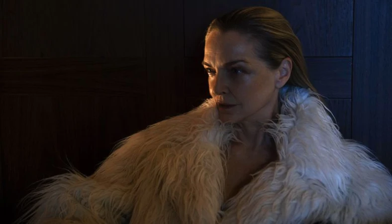 Portrait of an adult woman with blonde hair wearing a shaggy fur coat against a dark paneled wall, medium shot, neutral facial expression.