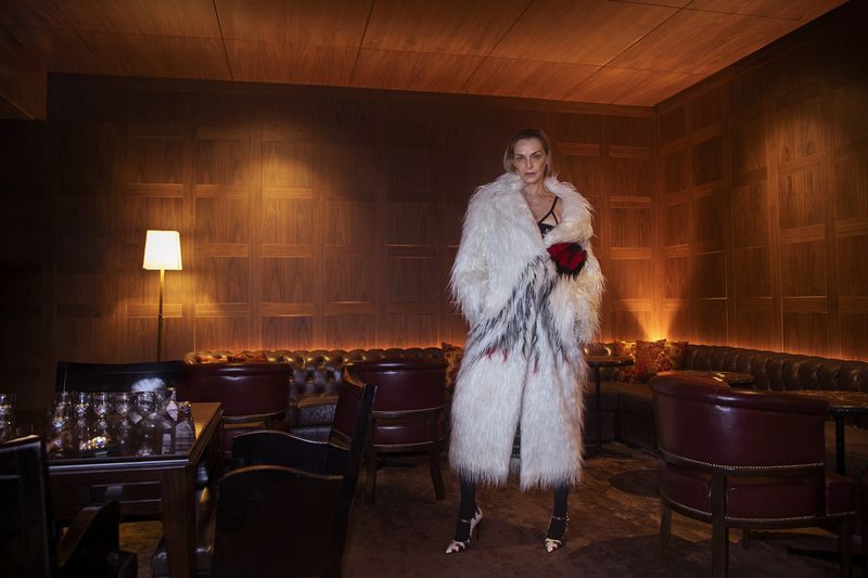Adult woman in a white faux fur coat with red and black accents poses in a dimly lit indoor setting with wood paneling and dark furniture.