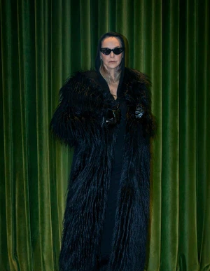 Simonetta Gianfelici in a long black shaggy coat, black hood, and dark sunglasses posing against green velvet curtains.