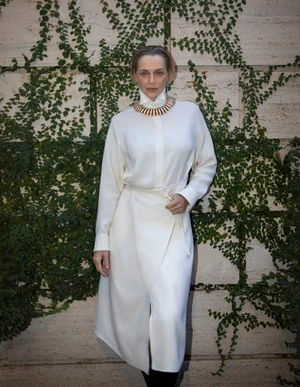 Simonetta Gianfelici poses in a long white belted dress and statement collar against an ivy-covered stone wall.