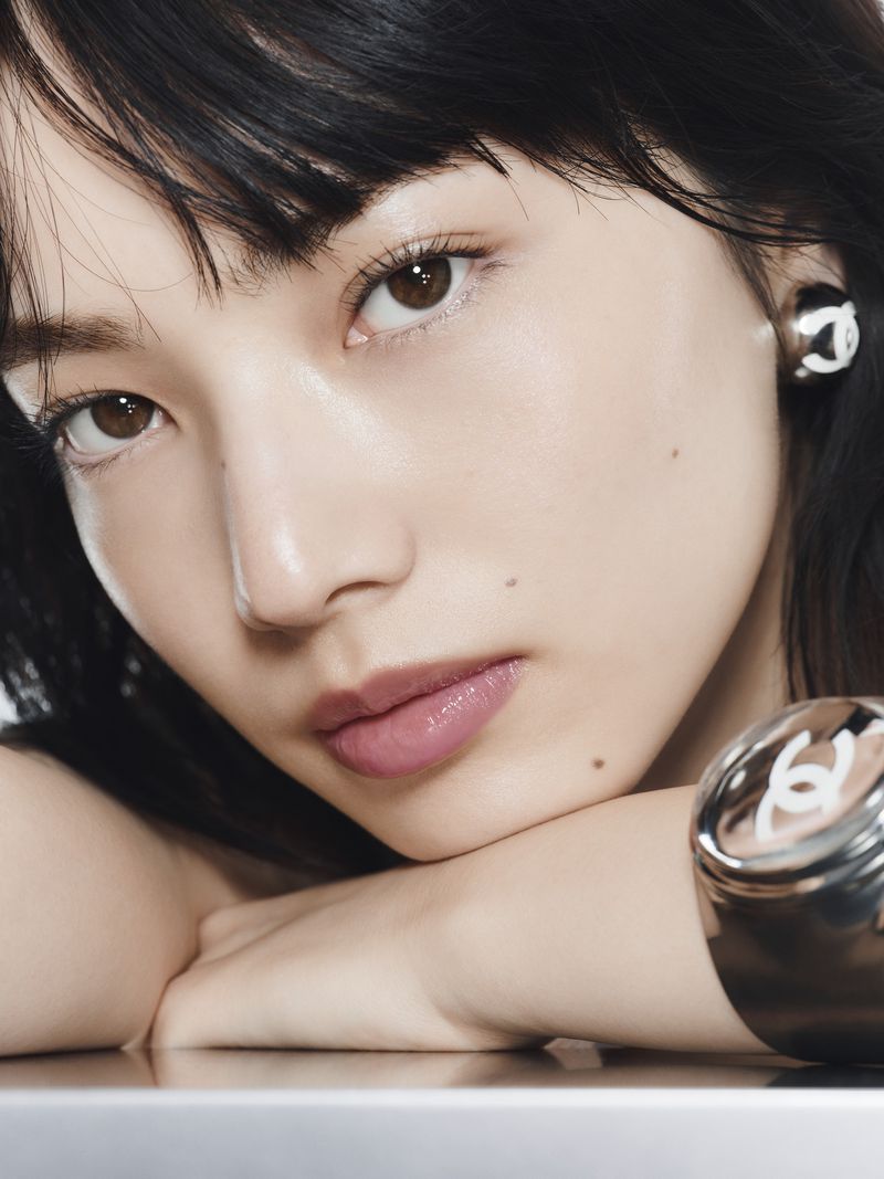 Close-up portrait of a young woman with dark hair, light skin, and brown eyes, wearing Chanel earrings and a silver cuff bracelet.