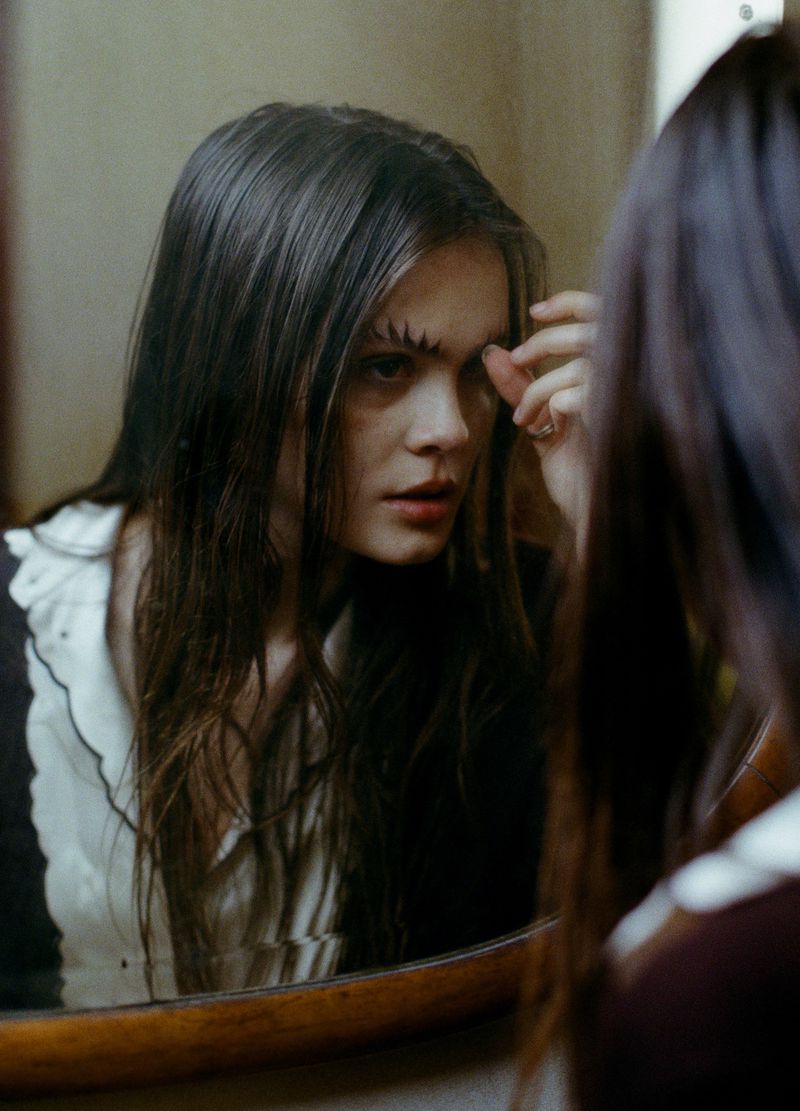 Young woman with long brown hair and unique eyebrow makeup gazes into a round mirror in an indoor setting, showcasing beauty and fashion.