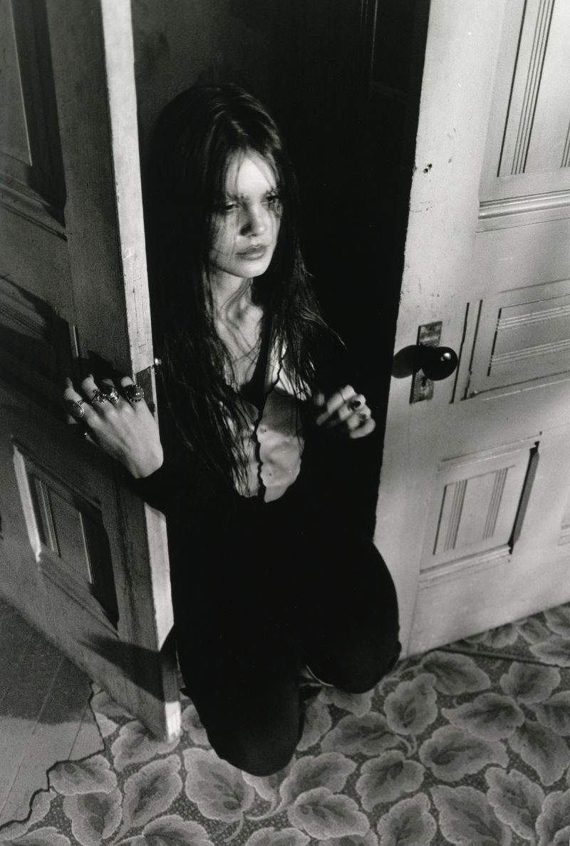 Monochrome portrait of a young woman with long dark hair standing in a doorway, looking away, wearing a dark top, and multiple rings.