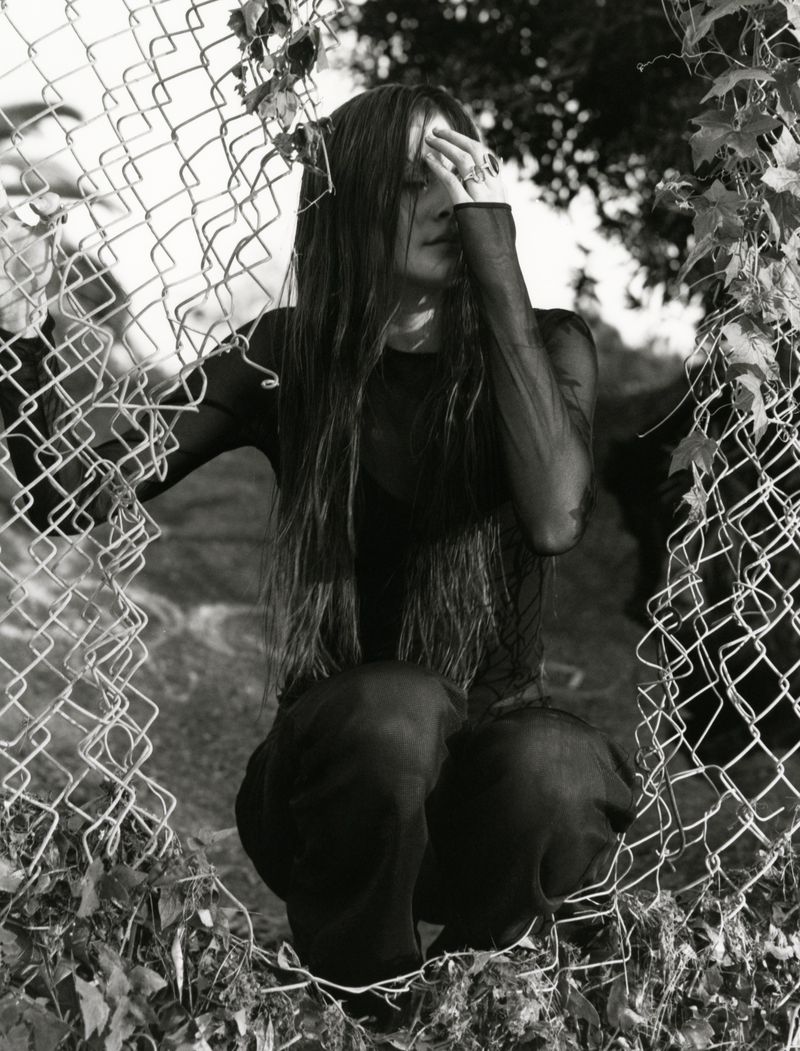 A woman with long hair and tattoos is kneeling behind a chain-link fence. She wears a dark, sheer top and matching pants. Black and white.