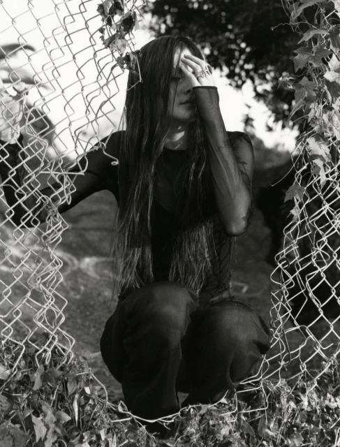 A woman with long hair and tattoos is kneeling behind a chain-link fence. She wears a dark, sheer top and matching pants. Black and white.