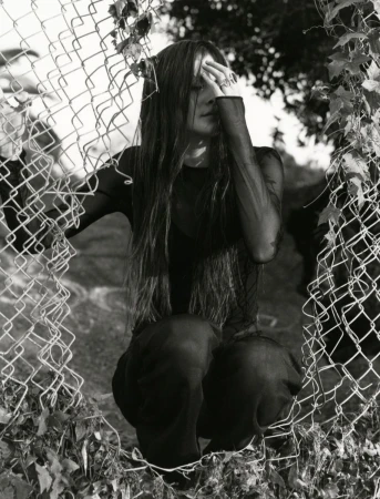 A woman with long hair and tattoos is kneeling behind a chain-link fence. She wears a dark, sheer top and matching pants. Black and white.