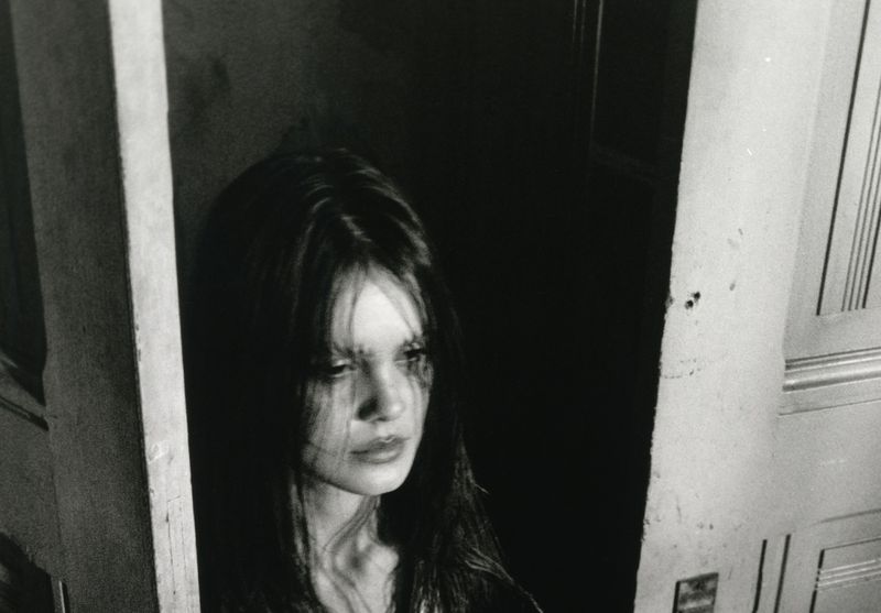 Black and white portrait of a woman with long dark hair, framed by a partially open doorway, with dramatic lighting.