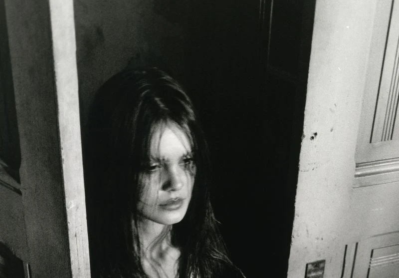 Black and white portrait of a woman with long dark hair, framed by a partially open doorway, with dramatic lighting.