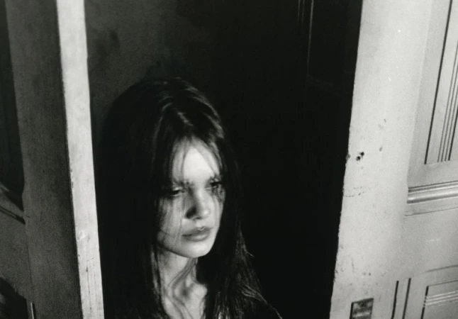 Black and white portrait of a woman with long dark hair, framed by a partially open doorway, with dramatic lighting.