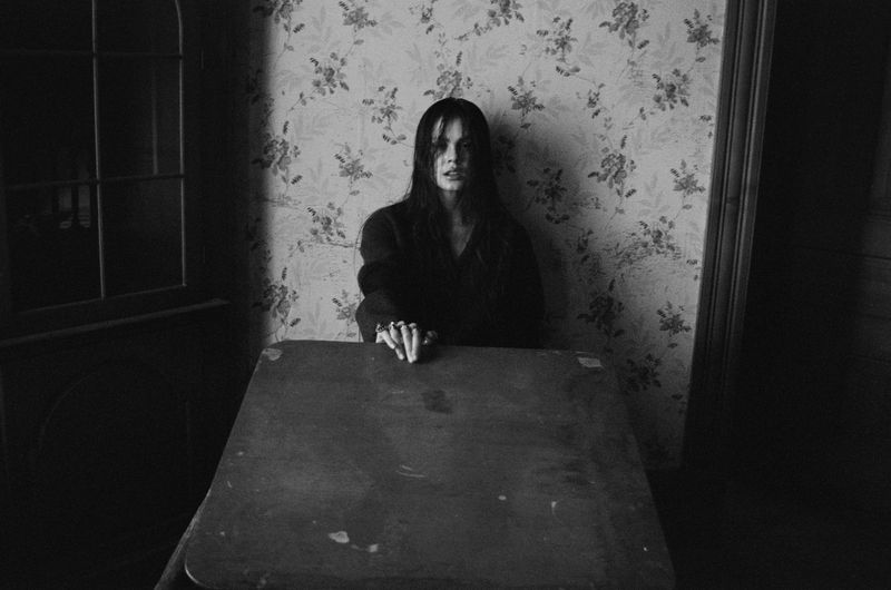Monochrome portrait of a young woman with long dark hair sitting at a wooden table indoors with floral wallpaper and a dark cabinet visible.