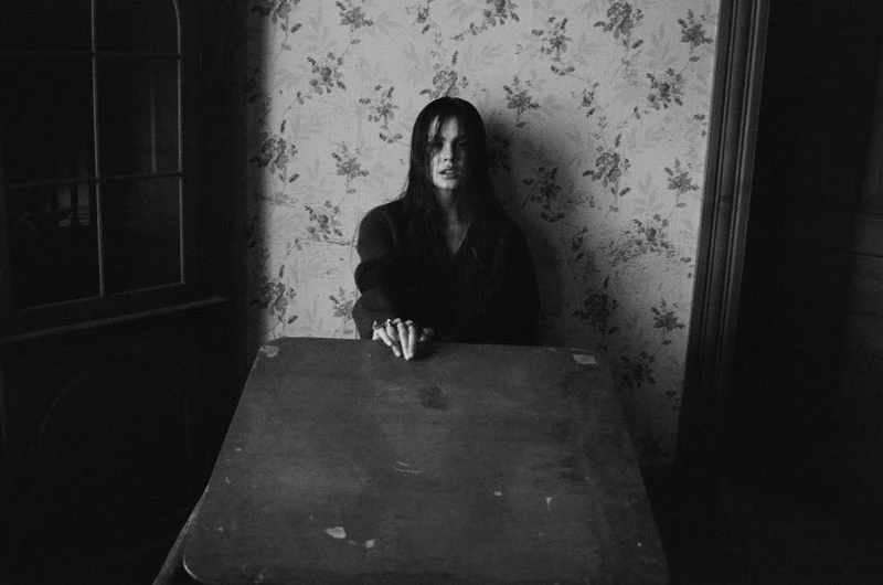 Monochrome portrait of a young woman with long dark hair sitting at a wooden table indoors with floral wallpaper and a dark cabinet visible.