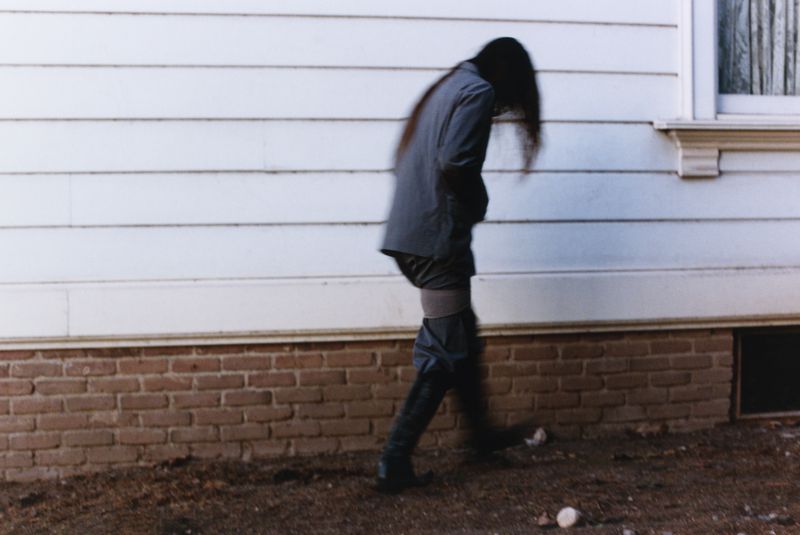 Person in blazer and boots walks in front of the white siding and brick foundation of a building, creating a blurred, dreamlike effect.