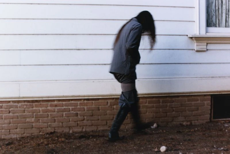 Person in blazer and boots walks in front of the white siding and brick foundation of a building, creating a blurred, dreamlike effect.