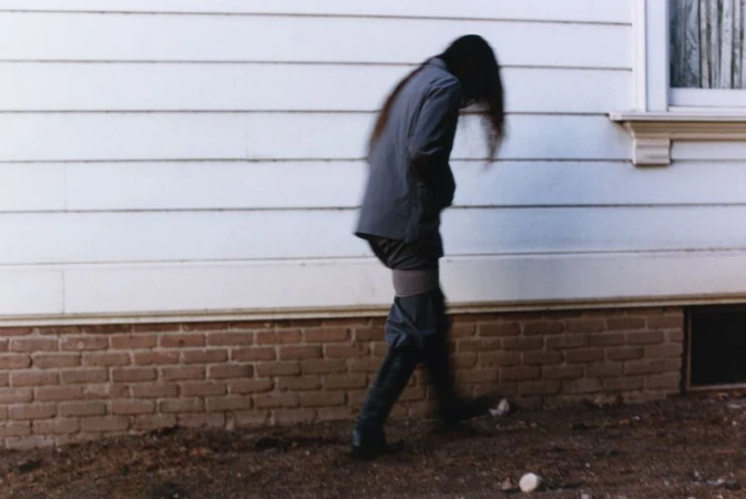 Person in blazer and boots walks in front of the white siding and brick foundation of a building, creating a blurred, dreamlike effect.