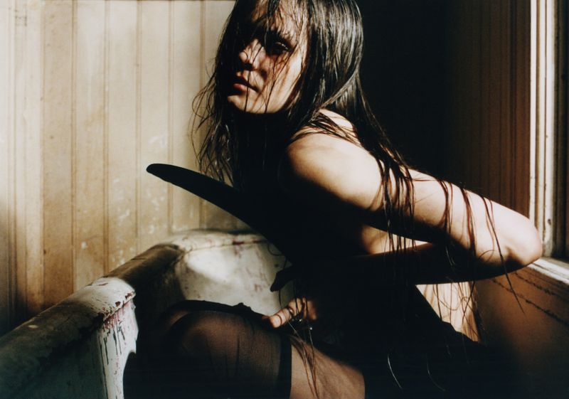 Dramatic photograph of a wet-haired, partially undressed adult female in a bathtub, emphasizing dark, moody lighting and a gritty aesthetic.