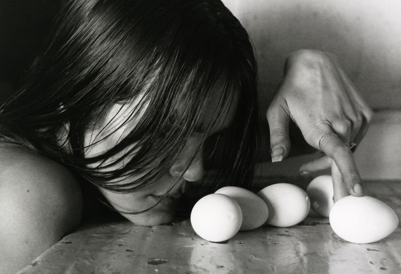 Black and white photograph of a woman with long, dark hair leaning over a table and touching one of several white eggs with her finger.