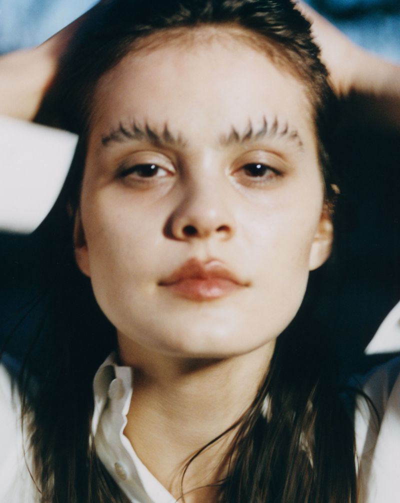 Close-up of a young woman with graphic eyebrows holding her hands behind her head, wearing a white shirt, against a blurred background.