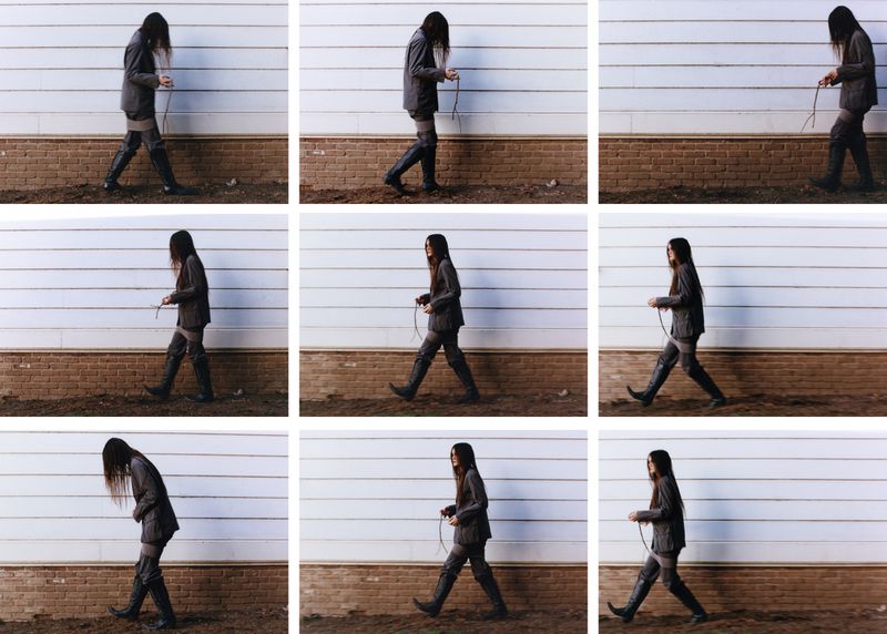 Adult in an asymmetrical, gray suit and dark boots, walking outdoors in a grid of nine frames against a white and brown wall.