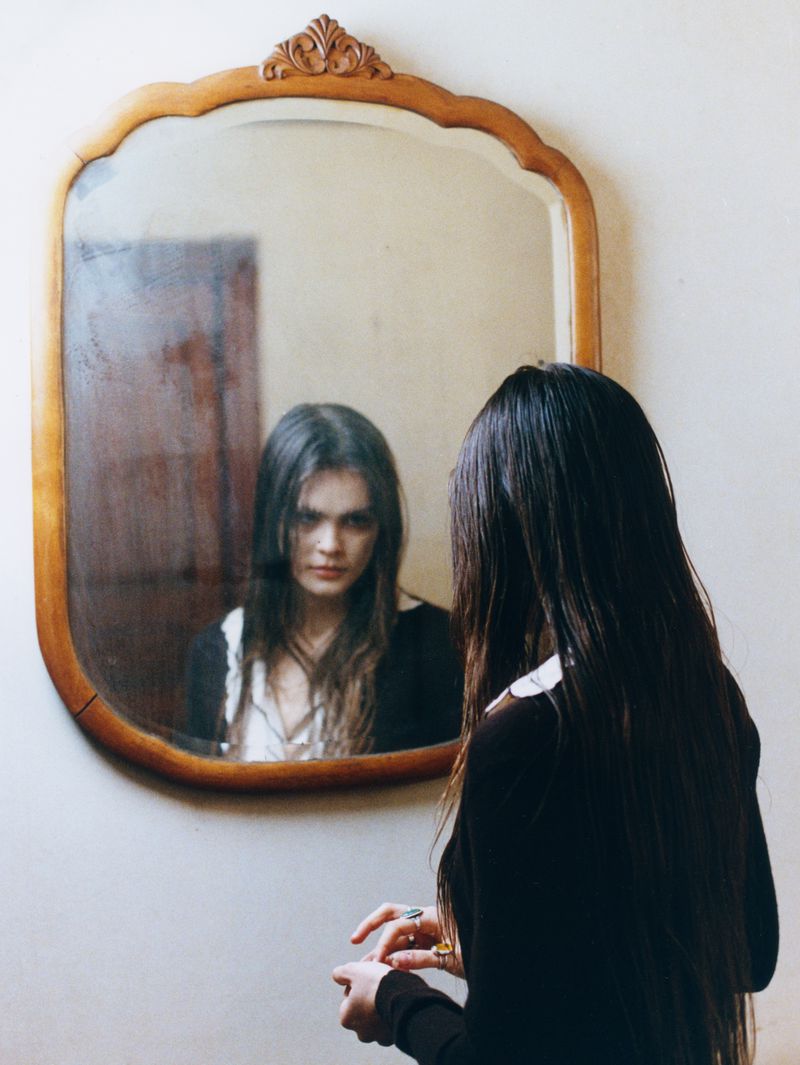 Woman in black dress gazes at reflection in antique mirror, light walls, indoor setting, long dark hair, rings on fingers.
