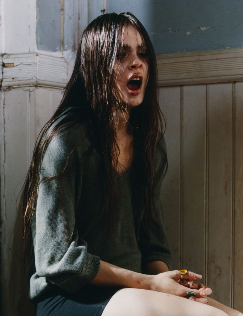 A young woman with dark hair, wearing a green sweater, sits with her mouth open in an old, dimly lit room with peeling white walls.
