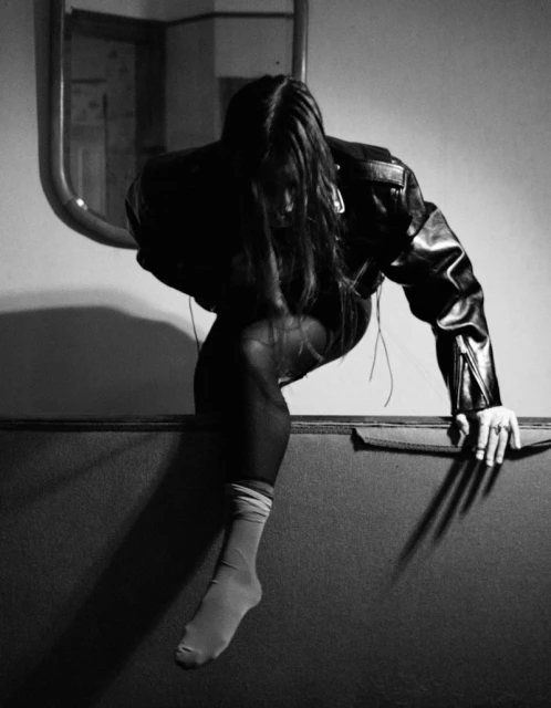 Person in leather jacket sitting on ledge with mirror and dark lighting creates dramatic, high-contrast, black and white portrait. 