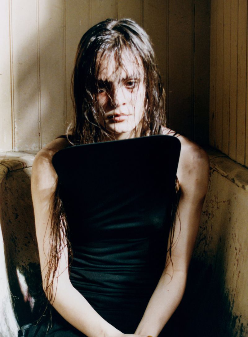A woman with long dark hair sitting in a bathtub. She is wearing a black sleeveless top and has a serious expression on her face.