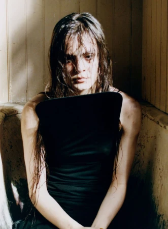 A woman with long dark hair sitting in a bathtub. She is wearing a black sleeveless top and has a serious expression on her face.