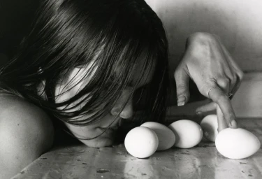 Sofia Isella resting her face on a textured surface beside white eggs in a black and white editorial portrait.