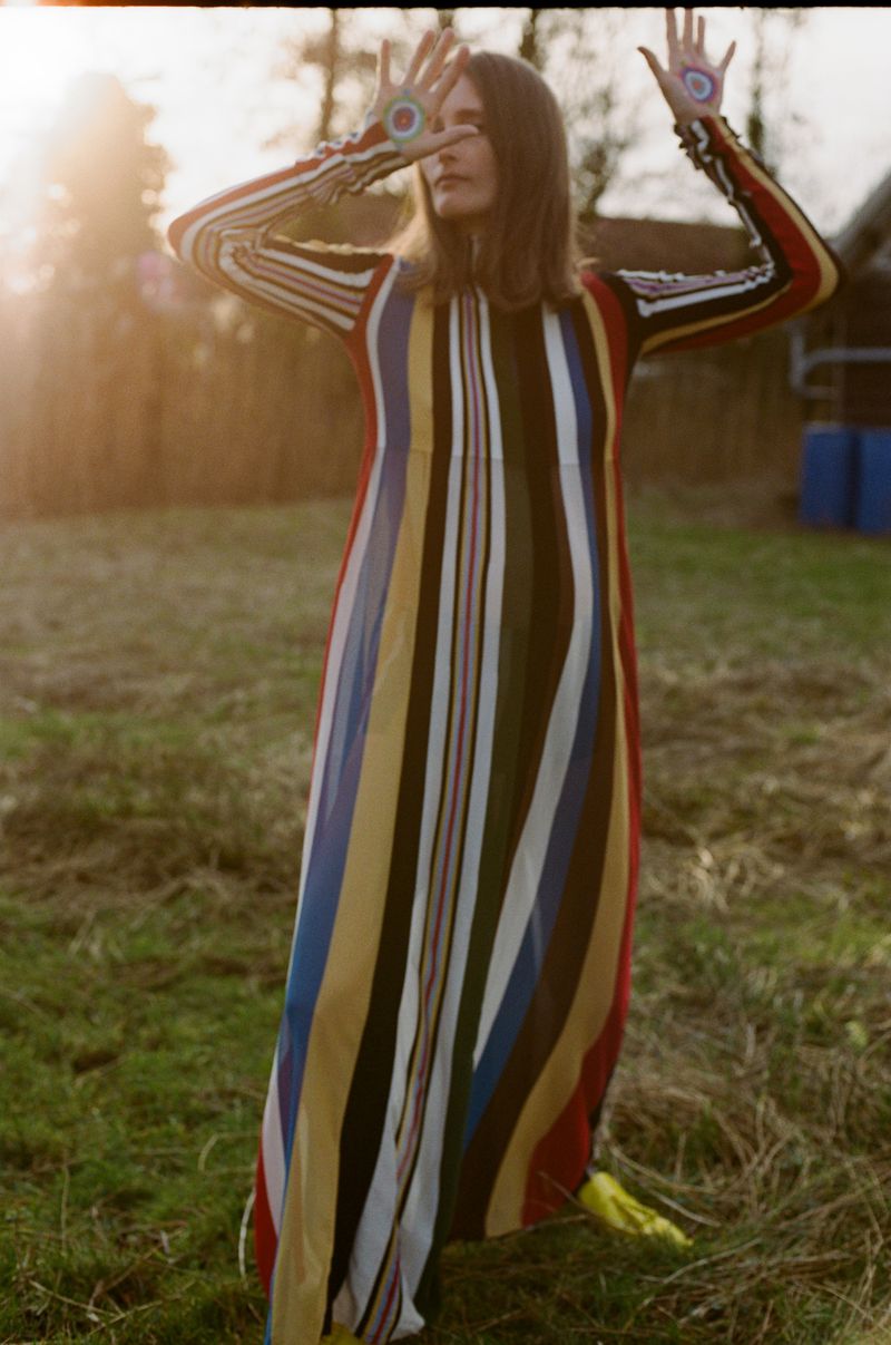 A woman with long brown hair stands in a grassy field, wearing a striped, long-sleeved dress and holding her hands up with designs on her palms.