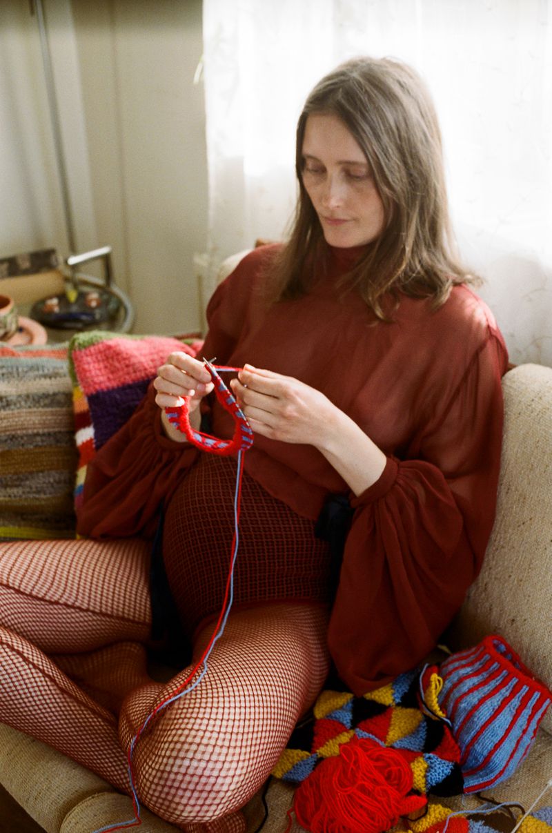 Pregnant woman in red fishnet tights sits knitting colorful yarn indoors. The photo features a cozy, domestic scene.