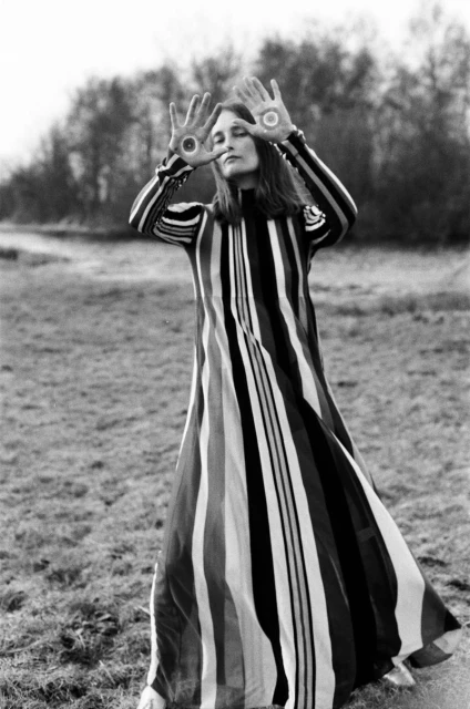 Full-length shot of an adult woman in a long striped dress with painted palms, standing outdoors in a grassy field under cloudy skies.