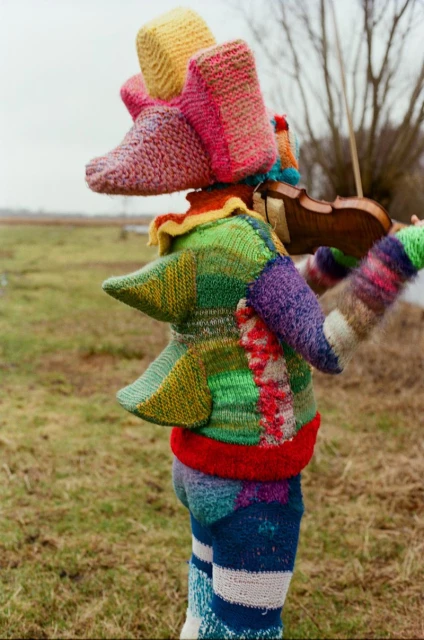 Unique photo of a knitted figure standing in a field, holding a violin, with a tree line and cloudy sky in the background, on an overcast day.