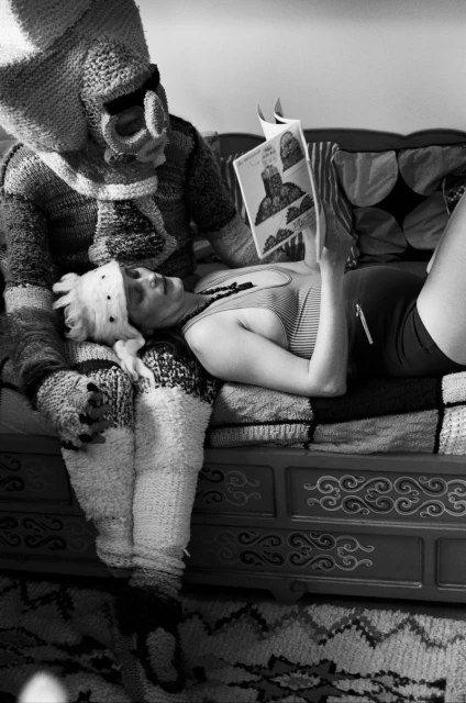 Black and white shot of a woman reclining while reading a book, with a person beside her wearing a knitted costume with large features.