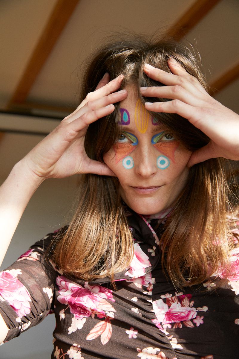 Close-up portrait of an adult female with colorful face paint framing her face with her hands, wearing a floral-patterned shirt.