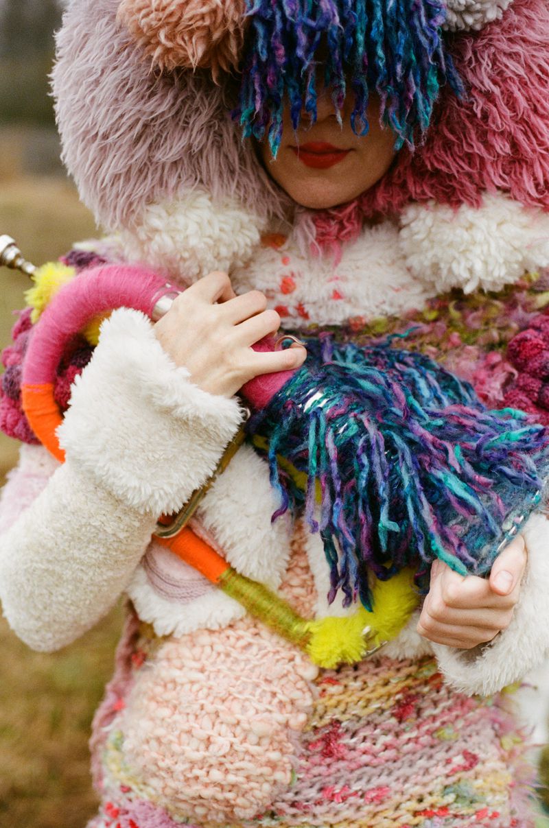 Woman in a colourful, textured coat with a furry hood, adorned with fringe, is standing outdoors with a blurred, grassy background.
