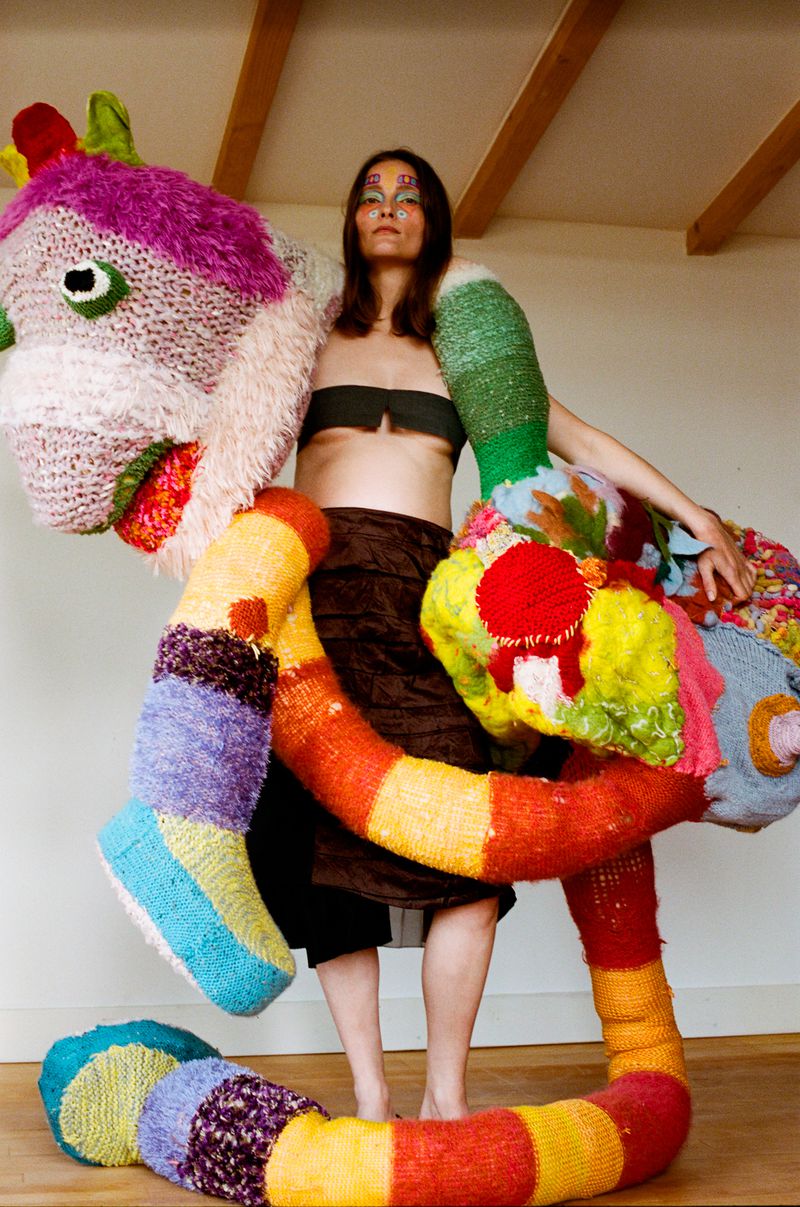 A woman poses indoors wearing a brown skirt and top, with a whimsical, large, knitted multi-colored animal-like creation around her.
