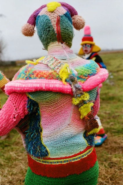 Two people in handmade, colorful knitwear stand outdoors. One is closer to the camera and the other is blurry in the background.