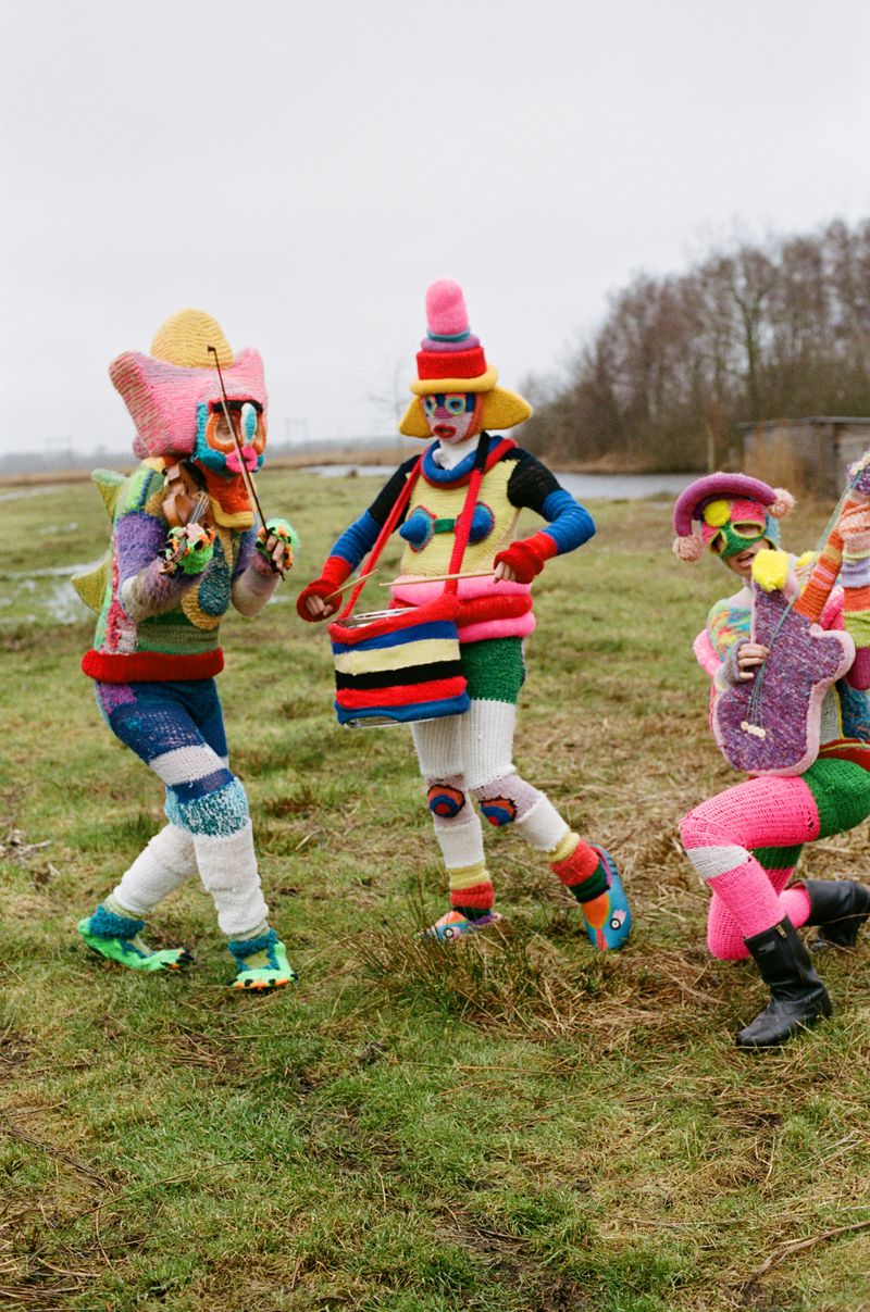 Three figures in colorful, knitted costumes play musical instruments on a grassy field under an overcast sky, creating a whimsical scene.