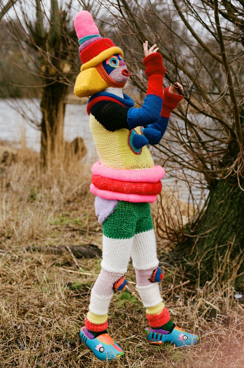 Person in knitted costume with colorful hat and patterned clothing stands in a field with bare trees and a body of water in the background.
