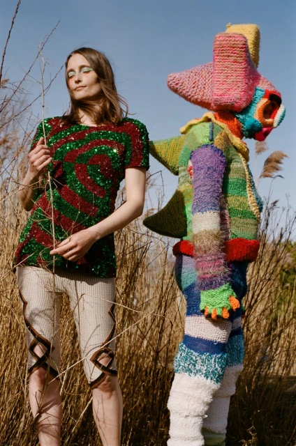 A woman in a sequined dress stands beside a knit sculpture in a field of tall grass under a clear sky in a fashion editorial.