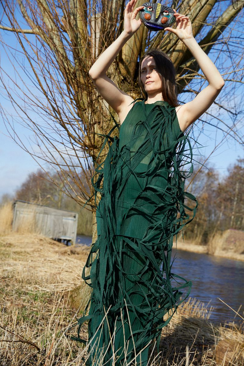 Adult female in green dress holding a colorful mask in a rural outdoor environment with bare trees and a body of water in the background.