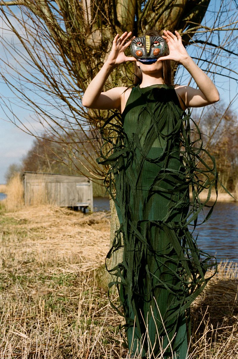 An adult female stands outdoors in a green dress, holding a tribal-style mask, with a river, bare trees, and a small wooden building in the background.