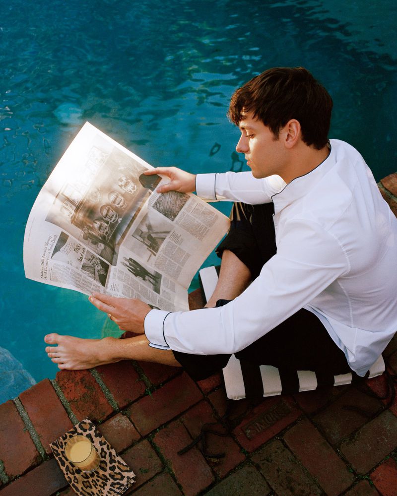 Young adult man in black and white pajamas sitting by the pool, reading a newspaper on a sunny day with a drink nearby.