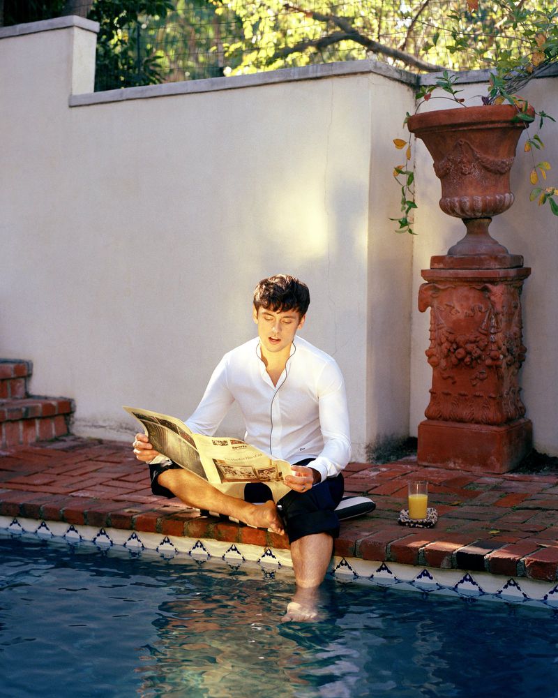 A relaxed man sits poolside reading a newspaper, with his feet in the water. There's a glass of orange juice and garden decor nearby.