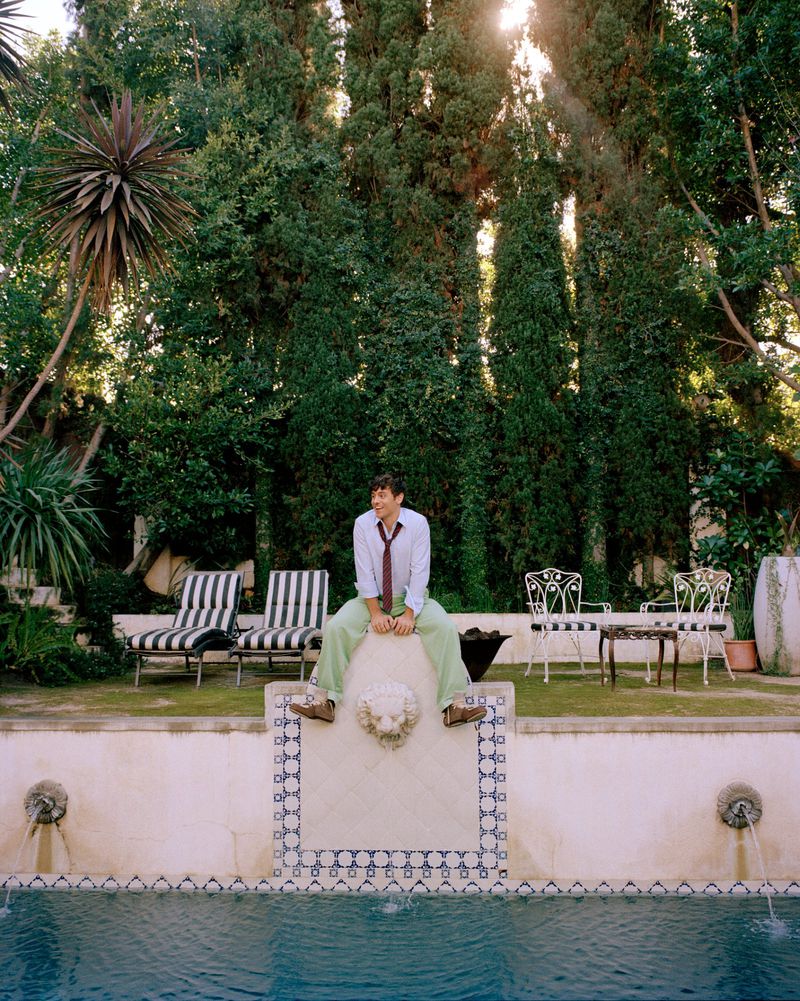 Man sitting on a ledge near a pool, wearing a white shirt, tie, and green pants with trees and lounge chairs in the background.
