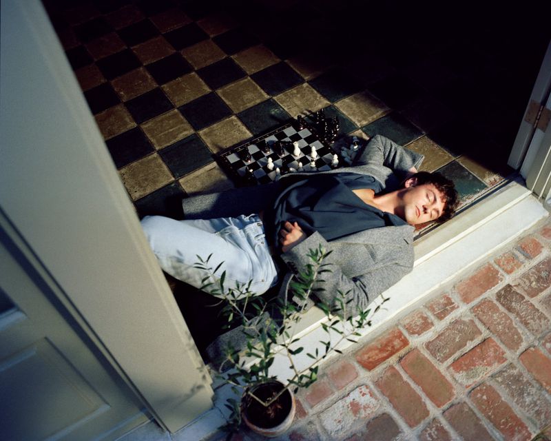 Young man in a grey jacket and blue jeans sleeps on a brick floor next to a chess board and potted plant in the corner of a room.