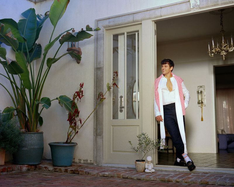 Stylish adult man posing in open doorway next to potted tropical plants in the courtyard of a bright, well-lit villa.