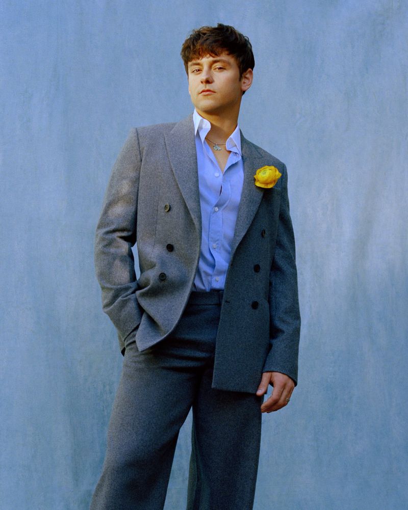 Young adult male in a grey suit and open blue shirt posing against a textured blue background with a yellow flower on the lapel.